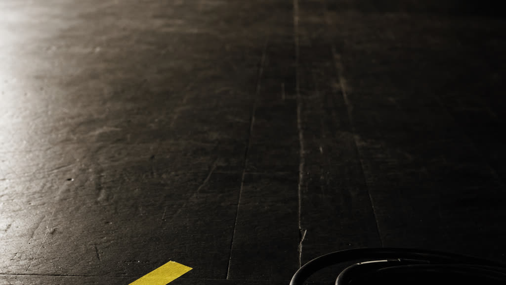 A strip of chrome yellow tape marking a spot on dark plywood staging, with a coiled cable beside it