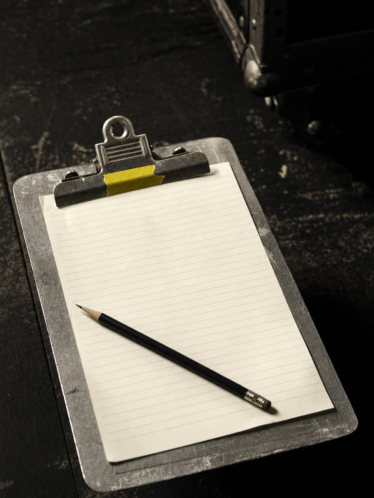 An aluminium clipboard on a dark surface holding a blank ruled sheet, a pencil across it and a yellow tape flag on the clip
